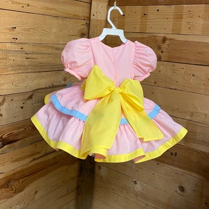 May include: A pink dress with a yellow bow and accents. The dress has short, puffy sleeves and a layered skirt with blue and yellow trim. It is hanging on a white hanger against a wooden background.