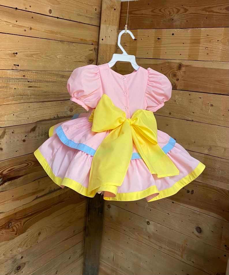 May include: A pink dress with a yellow bow and accents. The dress has short, puffy sleeves and a layered skirt with blue and yellow trim. It is hanging on a white hanger against a wooden background.