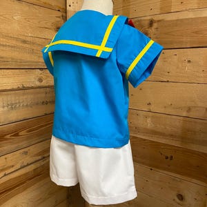 May include: A blue and white sailor outfit for a child. The shirt has yellow trim and a white collar. The shorts are white.
