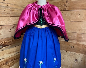 Puede incluir: Un vestido azul con flores bordadas y una capa de satén rosa con ribetes negros. El vestido tiene un cinturón dorado y un corpiño negro.