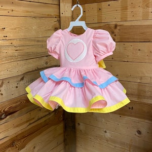 May include: A pink dress with puff sleeves and a tiered skirt. The bodice features a white circle with a pink heart. The skirt has blue and yellow trim. The dress is hanging on a white hanger against a wooden background.