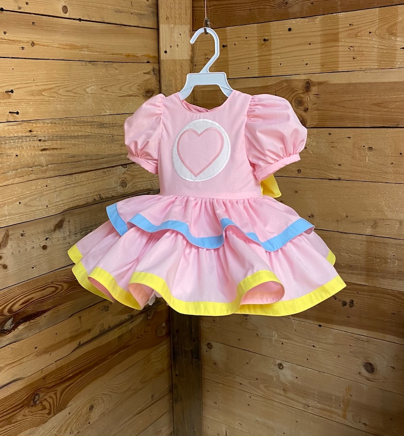 May include: A pink dress with puff sleeves and a tiered skirt. The bodice features a white circle with a pink heart. The skirt has blue and yellow trim. The dress is hanging on a white hanger against a wooden background.