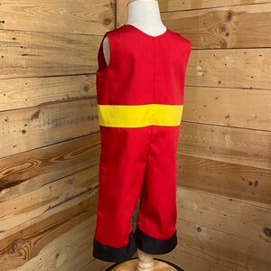 Peut inclure: Combinaison rouge avec une ceinture jaune et des poignets noirs, pr&eacute;sent&eacute;e sur un mannequin. La tenue sans manches a un col en V. Le costume est fait d'un tissu brillant.