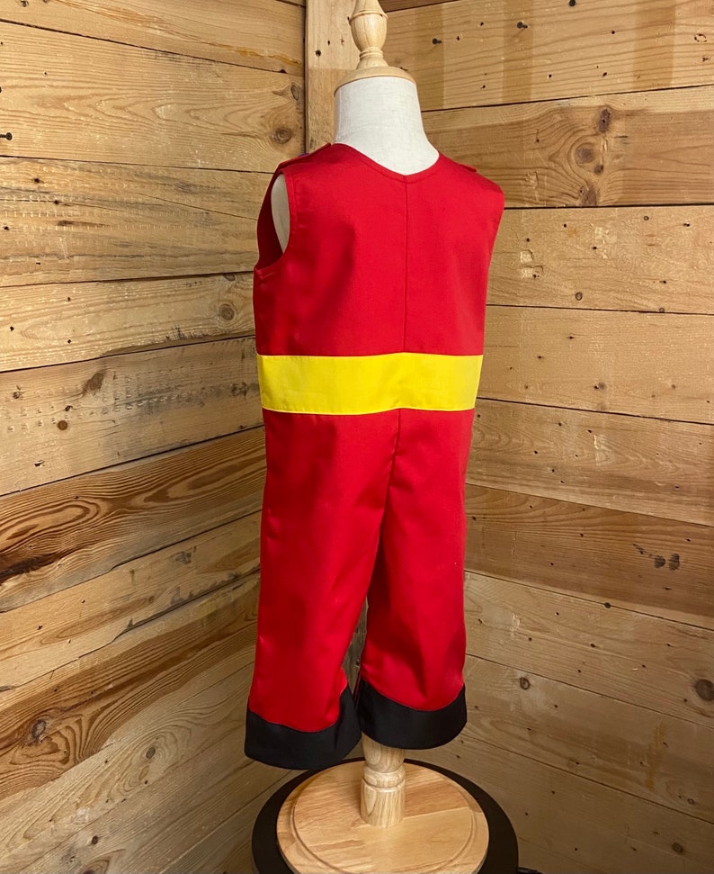 Peut inclure: Combinaison rouge avec une ceinture jaune et des poignets noirs, pr&eacute;sent&eacute;e sur un mannequin. La tenue sans manches a un col en V. Le costume est fait d'un tissu brillant.