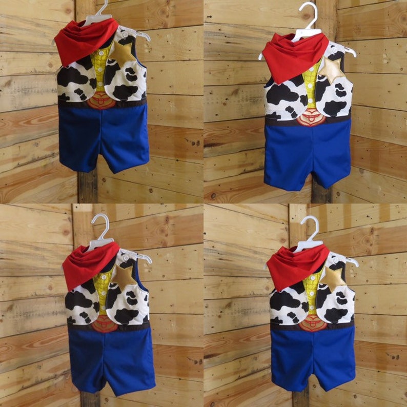 May include: A cowboy-themed costume for a baby or toddler, featuring a blue shorts set, a vest with a cow-print design, a yellow shirt, a red neckerchief, and a belt buckle detail. The costume is hanging on a white hanger.