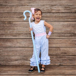 May include: A child's costume featuring a light blue jumpsuit with ruffled hems, a white lace bodice, and a pink waistband. The outfit includes a matching light blue shepherd's crook and a pink bow. The child is wearing black shoes.