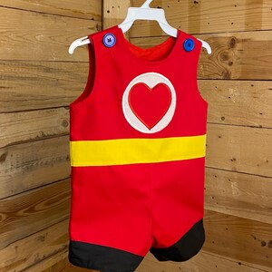 Puede incluir: Romper rojo con una franja amarilla y un emblema de coraz&oacute;n. El romper tiene botones azules en los hombros y detalles negros en los pantalones cortos. El conjunto est&aacute; colgado en una percha blanca sobre un fondo de madera.