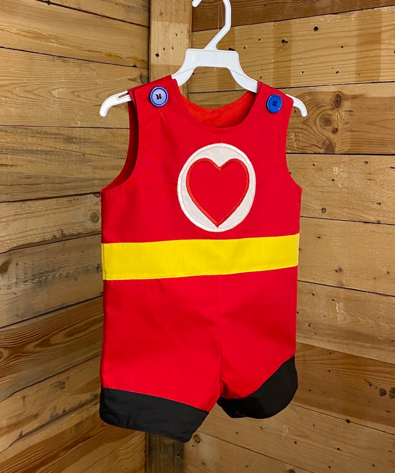 Puede incluir: Romper rojo con una franja amarilla y un emblema de coraz&oacute;n. El romper tiene botones azules en los hombros y detalles negros en los pantalones cortos. El conjunto est&aacute; colgado en una percha blanca sobre un fondo de madera.