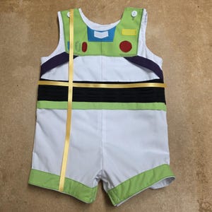 Puede incluir: Un pelele blanco y verde con el tema de Buzz Lightyear. La parte superior sin mangas presenta detalles verdes y azules, un c&iacute;rculo rojo y un rect&aacute;ngulo amarillo. Una banda negra y una cinta dorada detallan el frente. Los pantalones cortos tienen ribetes verdes.