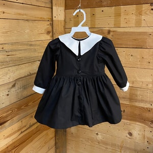 Pu&ograve; includere: Un abito nero con colletto bianco Peter Pan e maniche lunghe. L'abito ha una chiusura con bottoni sul davanti e una gonna a pieghe.