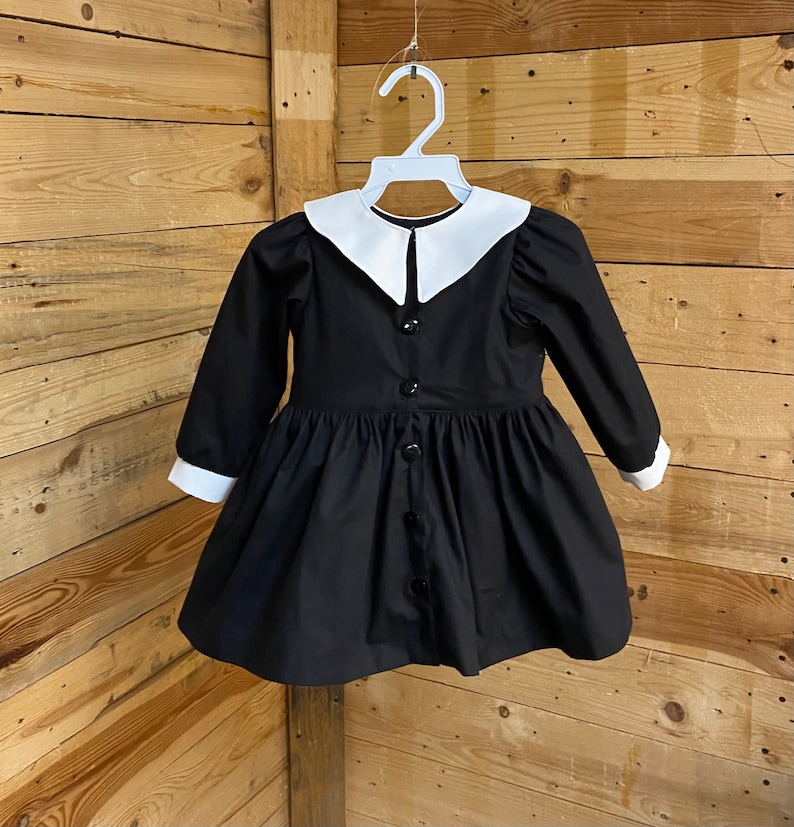 Pu&ograve; includere: Un abito nero con colletto bianco Peter Pan e maniche lunghe. L'abito ha una chiusura con bottoni sul davanti e una gonna a pieghe.