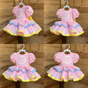 May include: Four pink dresses with puff sleeves and a heart design on the bodice are displayed on white hangers. Each dress has a layered skirt with blue and yellow trim. A yellow bow is attached to the back of each dress.