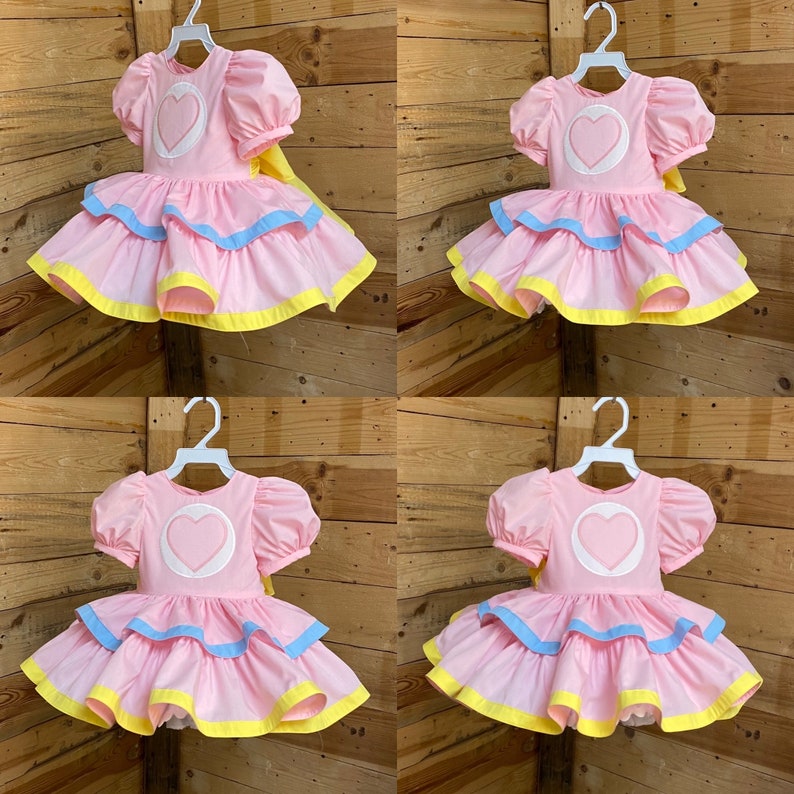 May include: Four pink dresses with puff sleeves and a heart design on the bodice are displayed on white hangers. Each dress has a layered skirt with blue and yellow trim. A yellow bow is attached to the back of each dress.