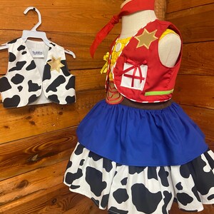 May include: A child's costume featuring a red vest with a gold star, a blue skirt, and a cow-print skirt layer. A matching cow-print vest hangs on a white hanger. The costume is displayed on a mannequin against a wood-paneled background.