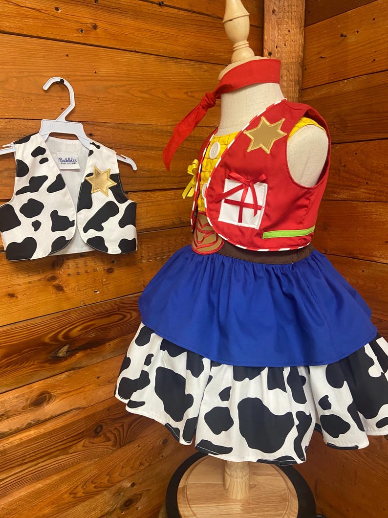 May include: A child's costume featuring a red vest with a gold star, a blue skirt, and a cow-print skirt layer. A matching cow-print vest hangs on a white hanger. The costume is displayed on a mannequin against a wood-paneled background.