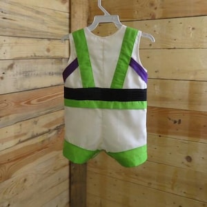 Puede incluir: Un disfraz blanco de Buzz Lightyear para ni&ntilde;o, colgado de una percha blanca. El disfraz tiene detalles verdes y morados, un cintur&oacute;n negro y ribetes verdes alrededor de las piernas.