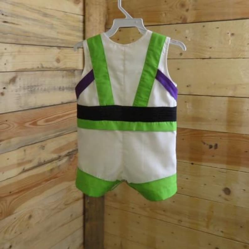 Puede incluir: Un disfraz blanco de Buzz Lightyear para ni&ntilde;o, colgado de una percha blanca. El disfraz tiene detalles verdes y morados, un cintur&oacute;n negro y ribetes verdes alrededor de las piernas.