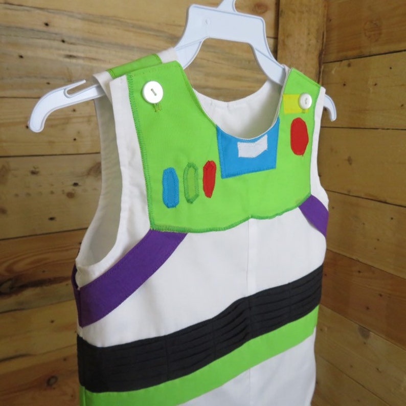 Puede incluir: Un vestido de disfraz blanco sin mangas con detalles en verde, morado y negro, que se asemeja a un uniforme de guardabosques espacial. El vestido presenta una pieza de pecho verde con detalles azules, rojos y amarillos, colgado de una percha blanca.
