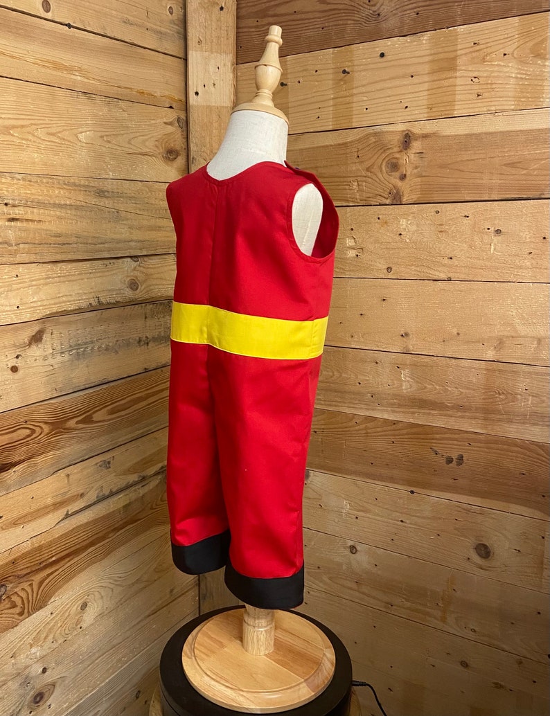Peut inclure: Un costume de pompier rouge et jaune pour enfants. Le costume comprend un haut et un pantalon rouges, avec une bande jaune sur la poitrine et une bordure noire aux chevilles. Le costume est pr&eacute;sent&eacute; sur un mannequin.