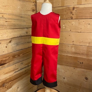 Peut inclure: Combinaison rouge et jaune pour enfant, pr&eacute;sent&eacute;e sur un mannequin en bois. La combinaison a une bande jaune au milieu et des poignets noirs aux chevilles. Le fond est un mur en bois.