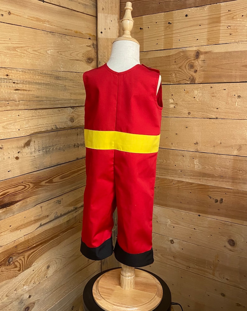 Peut inclure: Combinaison rouge et jaune pour enfant, pr&eacute;sent&eacute;e sur un mannequin en bois. La combinaison a une bande jaune au milieu et des poignets noirs aux chevilles. Le fond est un mur en bois.