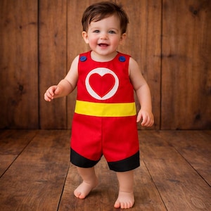 Puede incluir: Un pelele rojo para beb&eacute; con un coraz&oacute;n blanco, cinturilla amarilla y ribetes negros. El conjunto sin mangas tiene botones azules. Un ni&ntilde;o peque&ntilde;o sonriente lleva el pelele. El fondo es una superficie de madera.