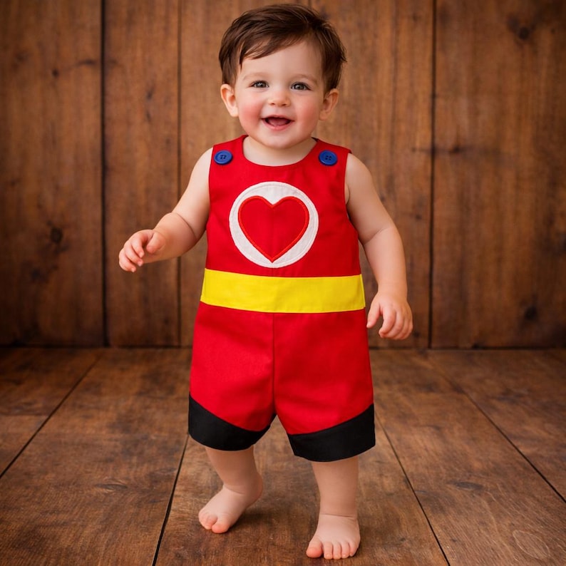 Puede incluir: Un pelele rojo para beb&eacute; con un coraz&oacute;n blanco, cinturilla amarilla y ribetes negros. El conjunto sin mangas tiene botones azules. Un ni&ntilde;o peque&ntilde;o sonriente lleva el pelele. El fondo es una superficie de madera.