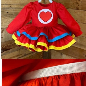 Pode incluir: Vestido vermelho com mangas compridas e saia rodada. O vestido tem um c&iacute;rculo branco com um cora&ccedil;&atilde;o vermelho no centro. A saia tem detalhes amarelos e azuis. O vestido tem uma cintura el&aacute;stica.