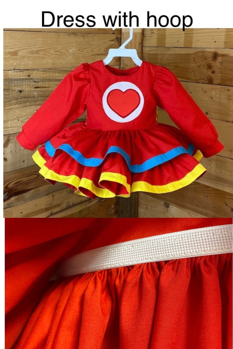 Pode incluir: Vestido vermelho com mangas compridas e saia rodada. O vestido tem um c&iacute;rculo branco com um cora&ccedil;&atilde;o vermelho no centro. A saia tem detalhes amarelos e azuis. O vestido tem uma cintura el&aacute;stica.