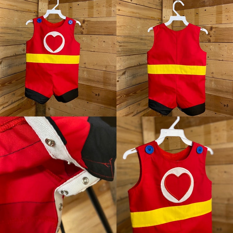 Puede incluir: Romper rojo con una franja amarilla y un dise&ntilde;o de coraz&oacute;n blanco. El romper tiene detalles negros en los pantalones cortos y botones azules en los hombros. La prenda se muestra en una percha blanca.