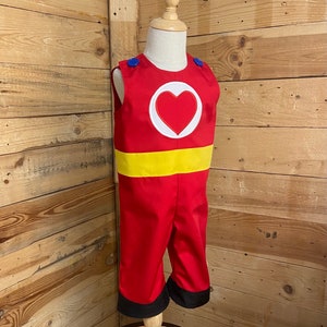 Peut inclure: Un costume de super-h&eacute;ros rouge et jaune pour enfant, avec un c&oelig;ur sur la poitrine. Le costume a des boutons bleus sur les &eacute;paules et des bordures noires sur le pantalon.