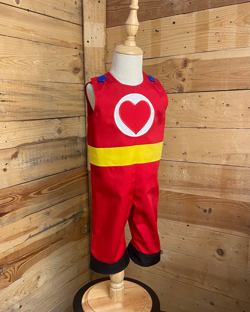 Peut inclure: Un costume de super-h&eacute;ros rouge et jaune pour enfant, avec un c&oelig;ur sur la poitrine. Le costume a des boutons bleus sur les &eacute;paules et des bordures noires sur le pantalon.