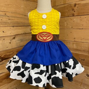 May include: A yellow and brown dress with a blue skirt and a cow print skirt. The top is yellow with a grid pattern and two white buttons. A brown belt with a red heart-shaped buckle is at the waist.