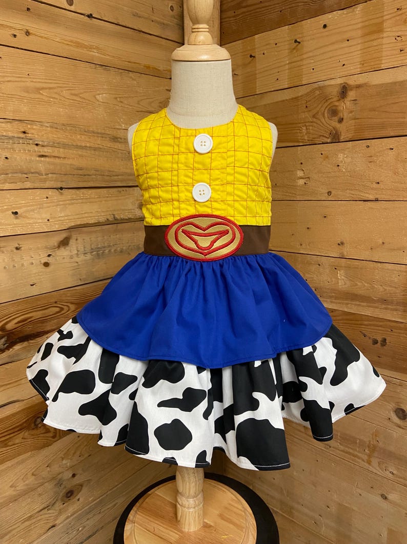 May include: A yellow and brown dress with a blue skirt and a cow print skirt. The top is yellow with a grid pattern and two white buttons. A brown belt with a red heart-shaped buckle is at the waist.