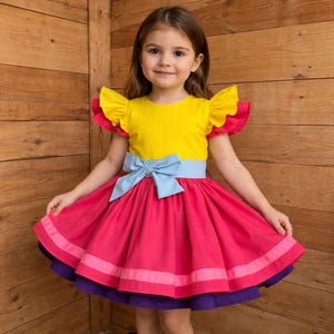 May include: A bright yellow and pink dress with a blue bow. The dress features ruffled sleeves and a full skirt with layers of pink and purple. The girl is smiling and standing in front of a wooden background.