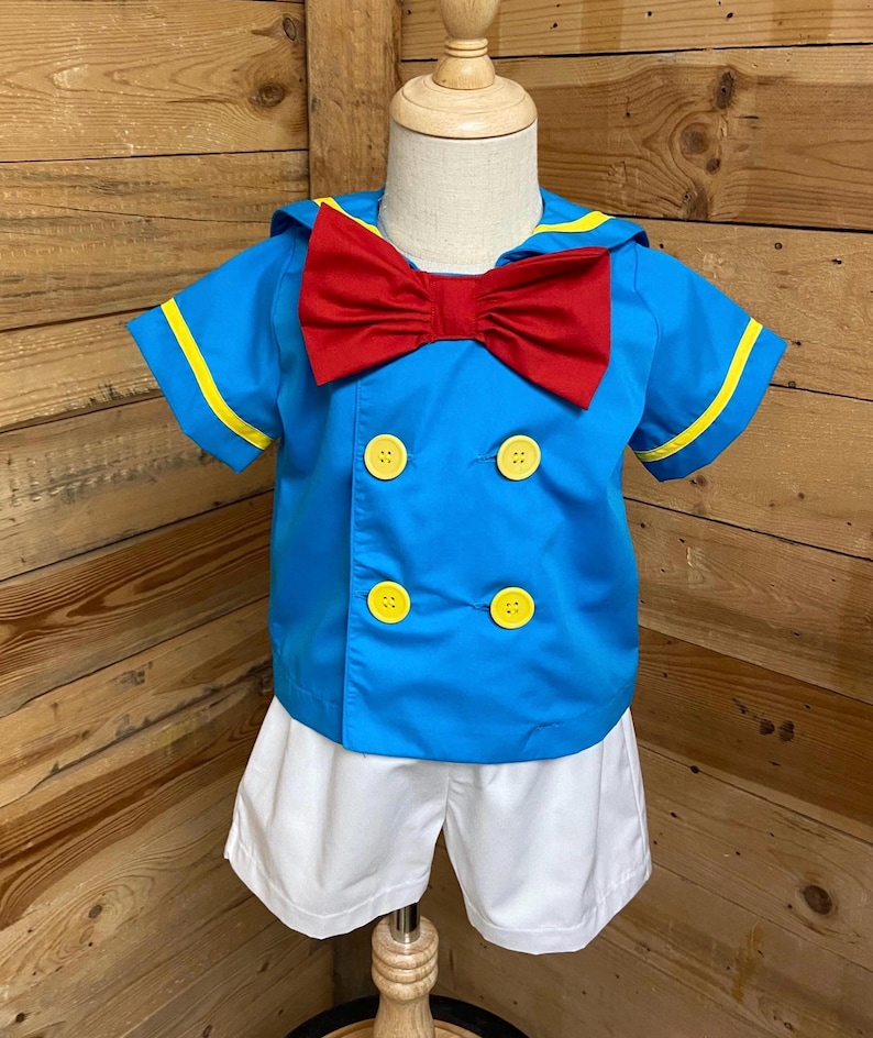 May include: A blue and white sailor-style outfit for a child. The outfit includes a short-sleeved shirt with a red bow tie, yellow trim, and four yellow buttons. The outfit also includes white shorts.