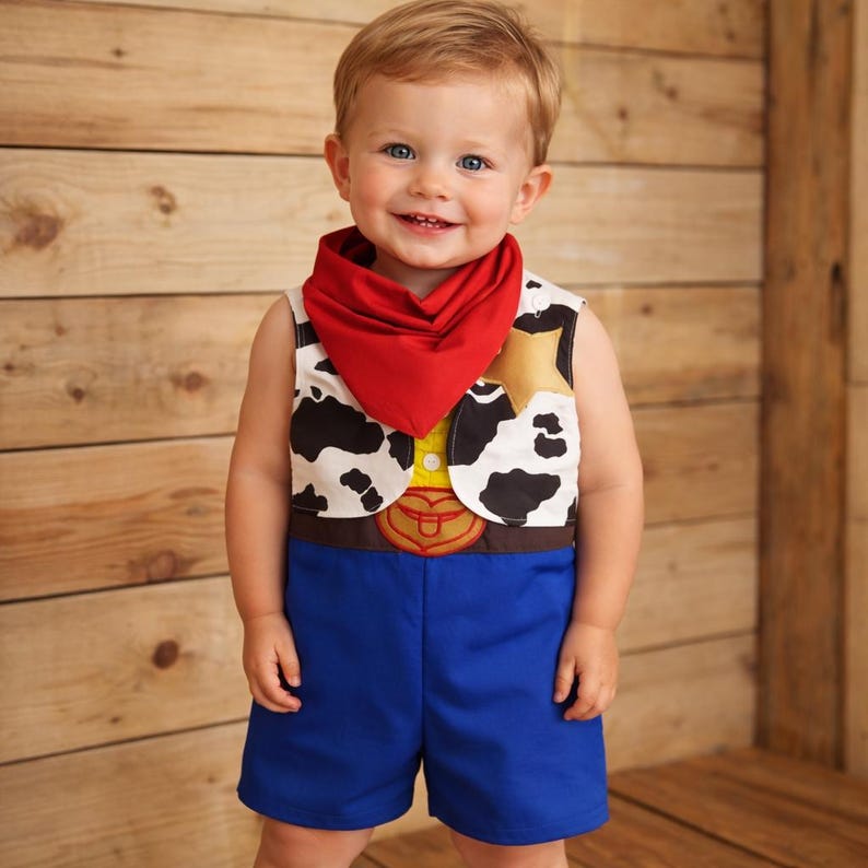 May include: A toddler wearing a cowboy costume, featuring a vest with a cowhide pattern, a red bandana, and blue shorts. The vest has a gold star and a brown belt buckle design. The child is smiling in front of a wooden background.