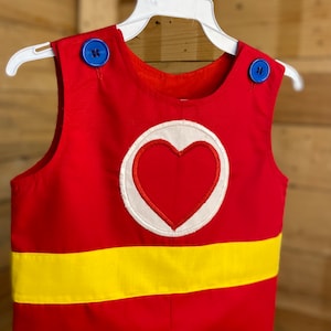 Puede incluir: Romper rojo con una franja amarilla y un emblema de coraz&oacute;n. El romper tiene botones azules en los hombros y est&aacute; colgado de una percha blanca. El emblema del coraz&oacute;n es rojo con un borde blanco.