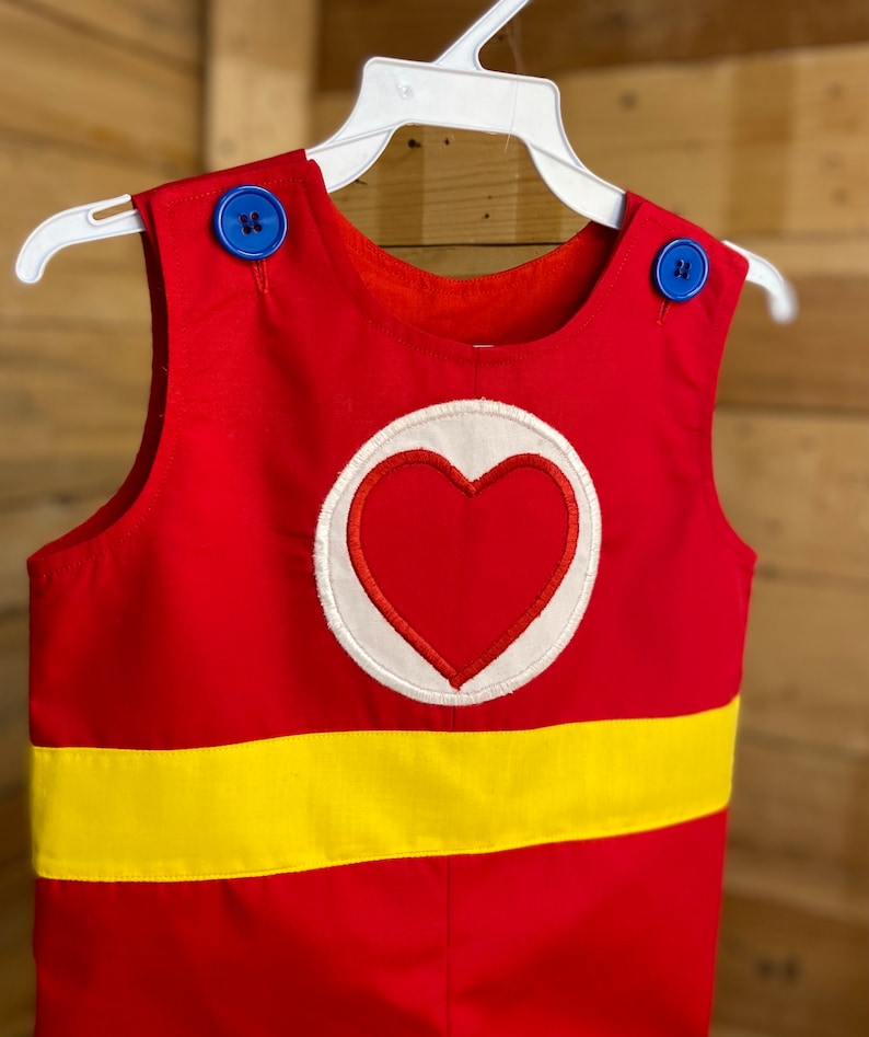 Puede incluir: Romper rojo con una franja amarilla y un emblema de coraz&oacute;n. El romper tiene botones azules en los hombros y est&aacute; colgado de una percha blanca. El emblema del coraz&oacute;n es rojo con un borde blanco.