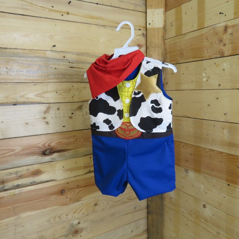 May include: A child's cowboy costume, featuring a blue romper with a yellow vest and a cow-print vest. A red scarf and a gold star complete the outfit. The costume is hanging on a white hanger against a wooden background.