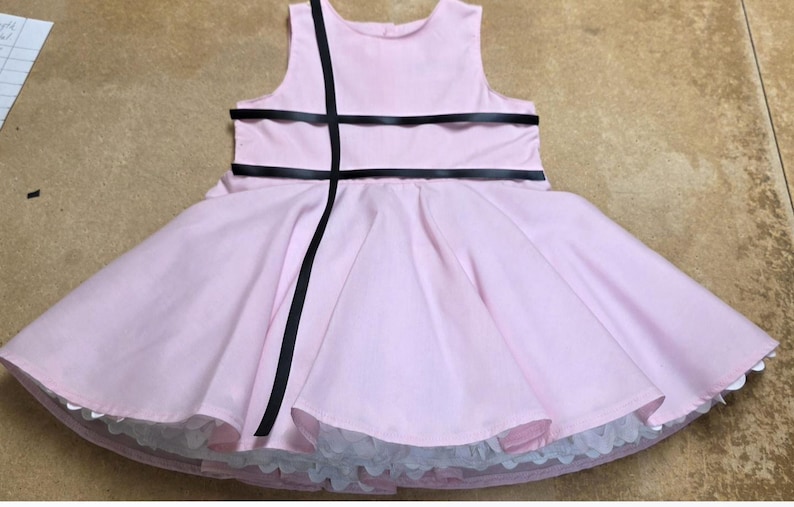 May include: A light pink dress with a full skirt and a sleeveless bodice. The dress is accented with black ribbon details across the bodice and down the front. The skirt has a white lace trim at the hem.
