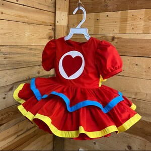 Pode incluir: Vestido vermelho com um cora&ccedil;&atilde;o branco e detalhes azuis e amarelos. O vestido tem uma saia rodada e est&aacute; pendurado em um cabide branco.