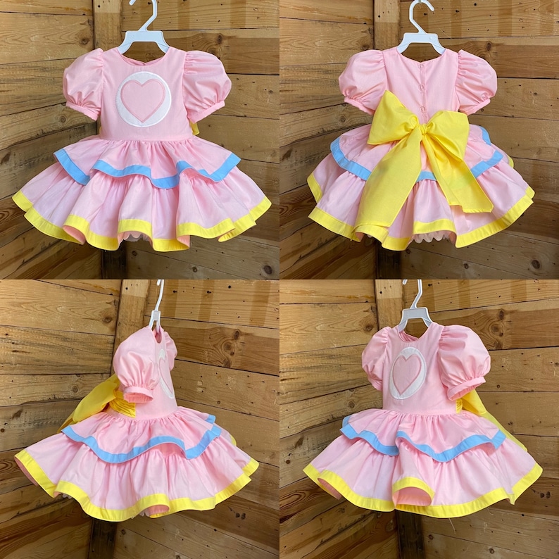 May include: A pink dress with a heart-shaped applique on the bodice and a large yellow bow. The dress features layered ruffles with blue and yellow trim, and puffy sleeves. The dress is hanging on a white hanger against a wooden background.