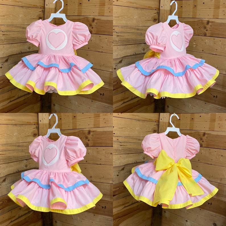 May include: Pink dress with a heart-shaped applique on the bodice and layered skirt. The dress features puff sleeves, blue and yellow trim, and a large yellow bow at the back. The dress is hanging on a white hanger.