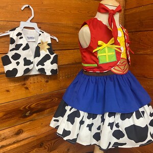 May include: A child's cowgirl costume featuring a red vest with a yellow and green design, a blue skirt, and a cow-print skirt. A matching cow-print vest hangs on a hanger. The costume is displayed on a mannequin against a wood background.