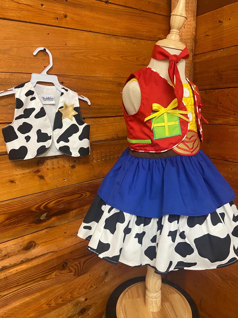 May include: A child's cowgirl costume featuring a red vest with a yellow and green design, a blue skirt, and a cow-print skirt. A matching cow-print vest hangs on a hanger. The costume is displayed on a mannequin against a wood background.