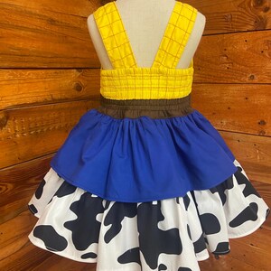 May include: A yellow and brown dress with blue and cow print skirt. The dress features yellow straps and a yellow bodice with a brown waistband. The skirt has a blue layer over a white layer with black cow spots.
