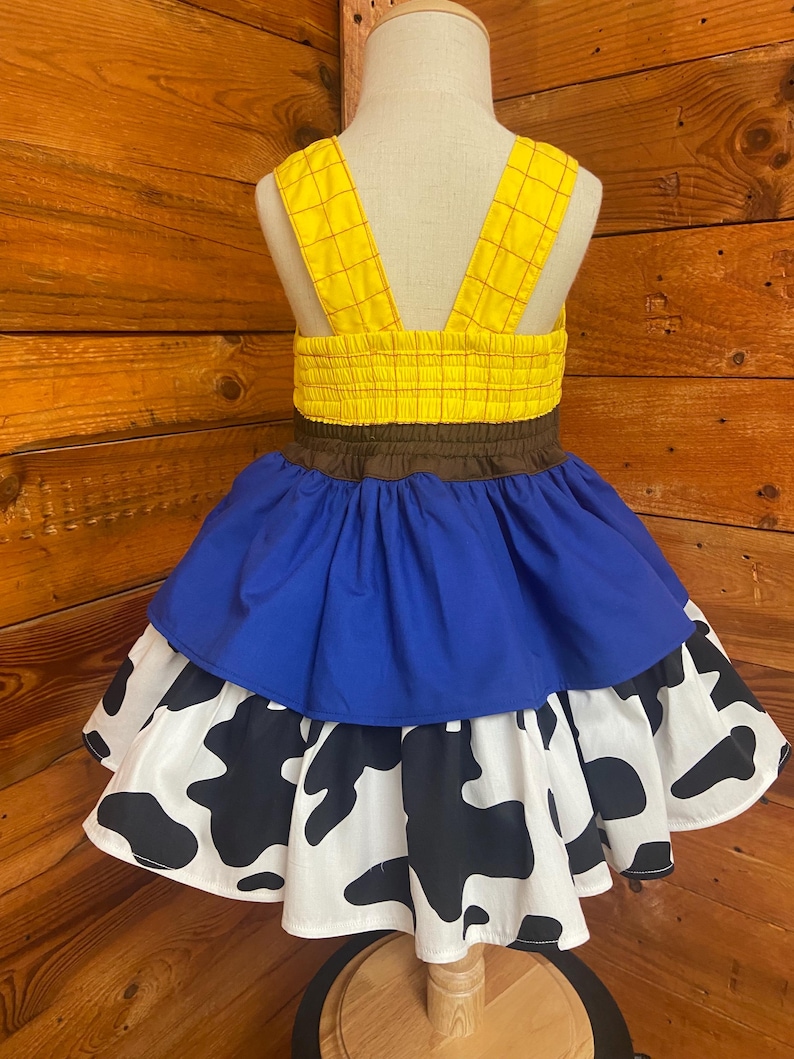 May include: A yellow and brown dress with blue and cow print skirt. The dress features yellow straps and a yellow bodice with a brown waistband. The skirt has a blue layer over a white layer with black cow spots.