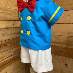 May include: A blue sailor shirt with yellow trim and a red bow tie. The shirt has three yellow buttons. The shirt is paired with white shorts.