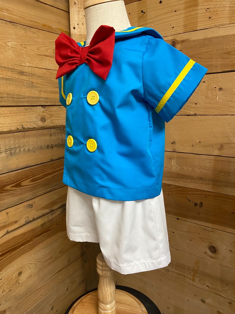 May include: A blue sailor shirt with yellow trim and a red bow tie. The shirt has three yellow buttons. The shirt is paired with white shorts.